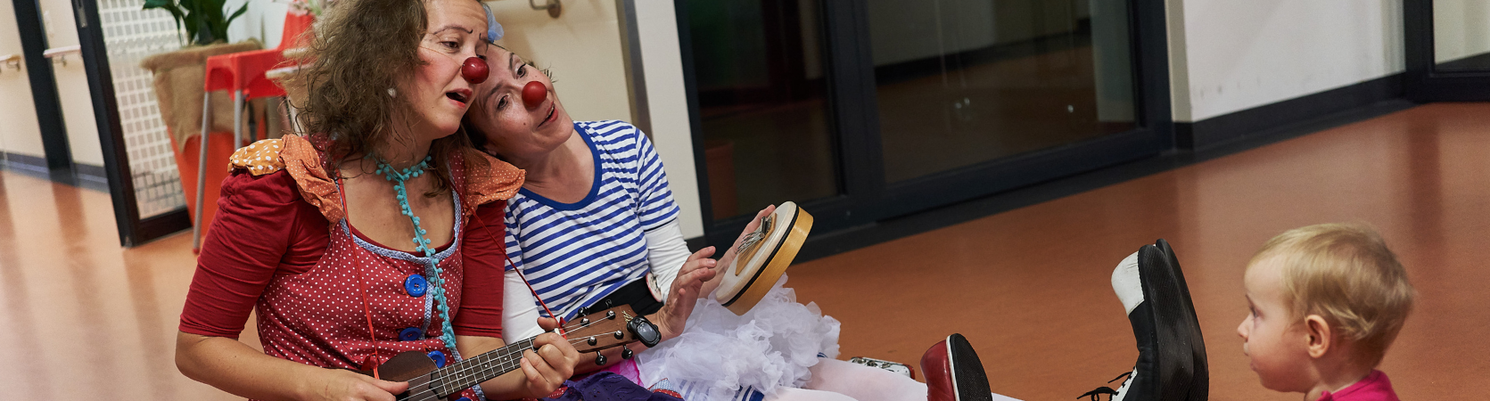 Zwei ROTE NASEN Clowns spielen einem Kleinkind ein Lied auf einer Ukulele vor.